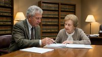 Senior Caucasian male attorney in suit consulting with elderly female client reviewing legal documents in traditional law office library setting.