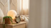 Cozy bedroom nightstand with vintage alarm clock, glowing lamp, green plant and knitted blanket creating warm atmospheric home decor for relaxation.