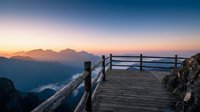 Wooden boardwalk with rustic railings extends toward mountain peaks at sunrise, overlooking misty valley and lake below for travel and nature backgrounds.