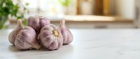 Fresh purple garlic bulbs with papery skin arranged on white marble countertop in bright kitchen setting. Suitable for food, cooking, and culinary content.