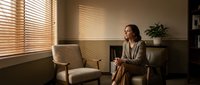 Professional businesswoman in beige suit sitting in modern office interior with wooden blinds creating warm natural lighting atmosphere.