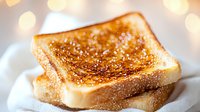 Freshly toasted white bread slice with golden-brown crispy surface and melted butter against soft bokeh background, close up view on white plate.