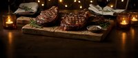 Grilled steaks on wooden cutting board with herbs and spices in warm candlelit restaurant setting for fine dining and culinary presentations.