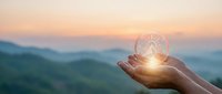 Hand holding glowing light bulb against mountain landscape at sunset symbolizing innovation, creativity, and bright ideas for business solutions.