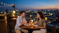 Romantic couple toasting with red wine on a rooftop restaurant at sunset, city skyline glowing in the background, warm string lights ambiance.