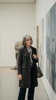 Mature woman with gray hair wearing dark blazer and shoulder bag viewing contemporary artwork in modern gallery space with white walls.
