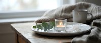 Cozy home interior with lit candle and eucalyptus branch on wooden tray beside window, creating peaceful hygge atmosphere for relaxation and mindfulness.