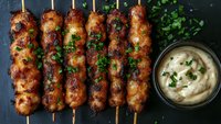 Grilled chicken kebabs on wooden skewers with fresh herbs and creamy dipping sauce on dark slate background for restaurant menu and cooking content.