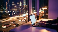 Professional working late on laptop by window with city lights bokeh background for remote work and urban lifestyle concepts.