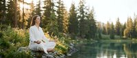 Young woman in white clothing practicing meditation by serene lake surrounded by evergreen forest during golden hour sunset creating peaceful wellness moment.