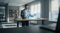 Professional businessman presenting holographic data visualization in modern office conference room with natural lighting and glass walls.
