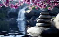 Balanced zen stones pyramid near waterfall with blooming pink cherry blossom branches above, reflecting in calm water surface against blurred nature background.