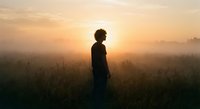 Young African American man standing in misty field during golden sunrise, silhouette against warm sky, peaceful morning meditation and self-reflection moment.