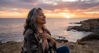 Mature woman with gray hair enjoying peaceful sunset moment by ocean coastline, expressing gratitude and mindfulness during golden hour meditation.