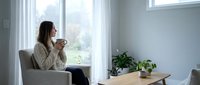 Young woman enjoying morning coffee in cozy armchair by window in bright modern living room with houseplants and natural light for relaxation and wellness.