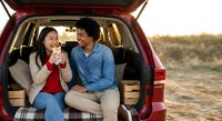 Young Hispanic couple enjoying coffee break during road trip adventure sitting in open car trunk at sunset outdoors together smiling and relaxing.
