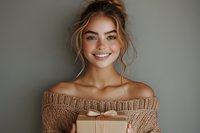 Young Caucasian woman with natural makeup and messy bun hairstyle smiling while holding kraft gift box, wearing off-shoulder knit sweater against gray background.