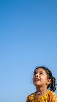 Young girl with dark hair and South Asian ethnicity wearing yellow and orange patterned clothing, looking upward with joy against clear blue sky background.