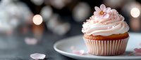 Pink frosted cupcake with delicate flower decoration on white plate surrounded by soft bokeh lights and scattered petals for bakery and celebration themes.