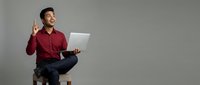 Young Asian male professional in maroon shirt sitting on stool with laptop, pointing upward with idea gesture for business, technology, and corporate concepts.