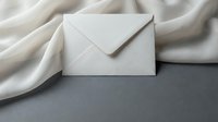 White envelope on gray surface with soft cream fabric drape creating elegant minimalist composition for correspondence and communication concepts.