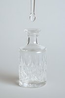 Crystal glass decanter with cut diamond pattern and liquid drop falling from dropper on light background, minimalist composition for cosmetic and spa concept.