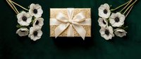 Elegant golden gift box with cream ribbon surrounded by white flowers on dark green background for luxury celebration occasions.