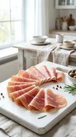 Thinly sliced prosciutto arranged on white marble board with rosemary and peppercorns in bright kitchen setting for gourmet appetizer presentation.