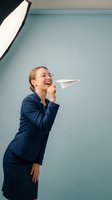 Young Caucasian businesswoman in navy blazer and glasses throwing paper airplane, expressing joy and innovation in professional setting.