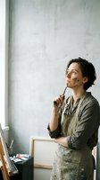 Young male artist with dark hair and East Asian ethnicity, wearing olive-green shirt and paint-stained apron, holding paintbrush thoughtfully while gazing upward in creative studio space with white wall.