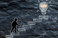 Man climbing stairs drawn with chalk on dark stone texture leading to glowing light bulb, representing growth, innovation and reaching goals in business concept.