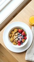 Overhead view of a white bowl filled with golden cereal, fresh raspberries, and blueberries on a light wooden table with orange juice, perfect for breakfast and healthy eating content.