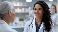 Young female Hispanic doctor with stethoscope and white coat smiling warmly while consulting senior patient during medical appointment in modern clinic office setting.