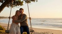 Young Caucasian couple embracing on wooden tree swing at golden hour beach sunset, romantic moment by ocean waves with warm lighting for wedding concepts.