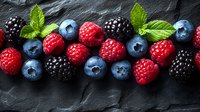 Fresh mixed berries arranged in row on dark slate background with mint leaves, including ripe raspberries, blackberries and blueberries for food design.