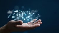 Open hand displaying glowing digital business icons including graphs, charts, magnifying glass, and data symbols on dark blue background for analytics and technology concepts.