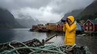 Person wearing yellow rain jacket photographing scenic fjord landscape with traditional red wooden cabins, fishing nets, and moody mountain scenery, suitable for travel, nature, and lifestyle content.