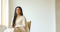 Young woman with long braided hair and South Asian ethnicity, wearing an elegant cream-colored linen shirt, seated on a wooden chair in a bright, minimalist interior space with natural window lighting.
