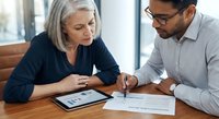 Senior Caucasian businesswoman and young Hispanic businessman collaborating on financial documents and digital tablet in modern office meeting room.