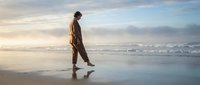Young man walking alone on wet beach sand during golden hour sunset with gentle waves and cloudy sky creating peaceful solitude moment for wellness concepts.