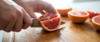 Close-up of hand slicing fresh grapefruit on wooden cutting board with knife, food preparation and cooking ingredient for recipes and culinary content.