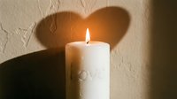 Burning white pillar candle with love inscription casting heart-shaped shadow on textured wall, romantic Valentine's Day atmosphere.