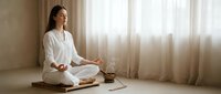 Young Asian woman in white clothing practicing meditation in peaceful indoor setting with natural light streaming through curtains.