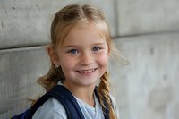 Cheerful young student with braided hair and blue eyes wearing backpack smiles brightly against concrete wall background, expressing joy and confidence on first day of school.