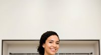 Young woman with dark hair and olive complexion smiling at camera in modern interior setting, suitable for business, lifestyle, and corporate website design projects.