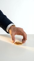 Businessman in dark suit and white cuff reaching toward gold coin on white surface, symbolizing wealth, investment, and financial opportunity for business concepts.