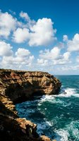 Dramatic rocky coastal cliffs with turquoise ocean waves and white fluffy clouds in bright blue sky, perfect for travel and nature backgrounds.