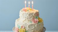 Elegant two-tier birthday cake with white frosting, colorful sugar roses and three lit candles on blue background for celebration events.