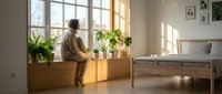 Young man with curly hair and medium brown skin tone tending to potted plants on wooden shelf by sunlit window in minimalist home interior, lifestyle and plant care concept.