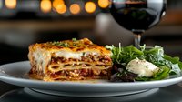 Layered pasta dish with meat sauce and melted cheese served on white plate with fresh greens and red wine for fine dining and restaurant menus.